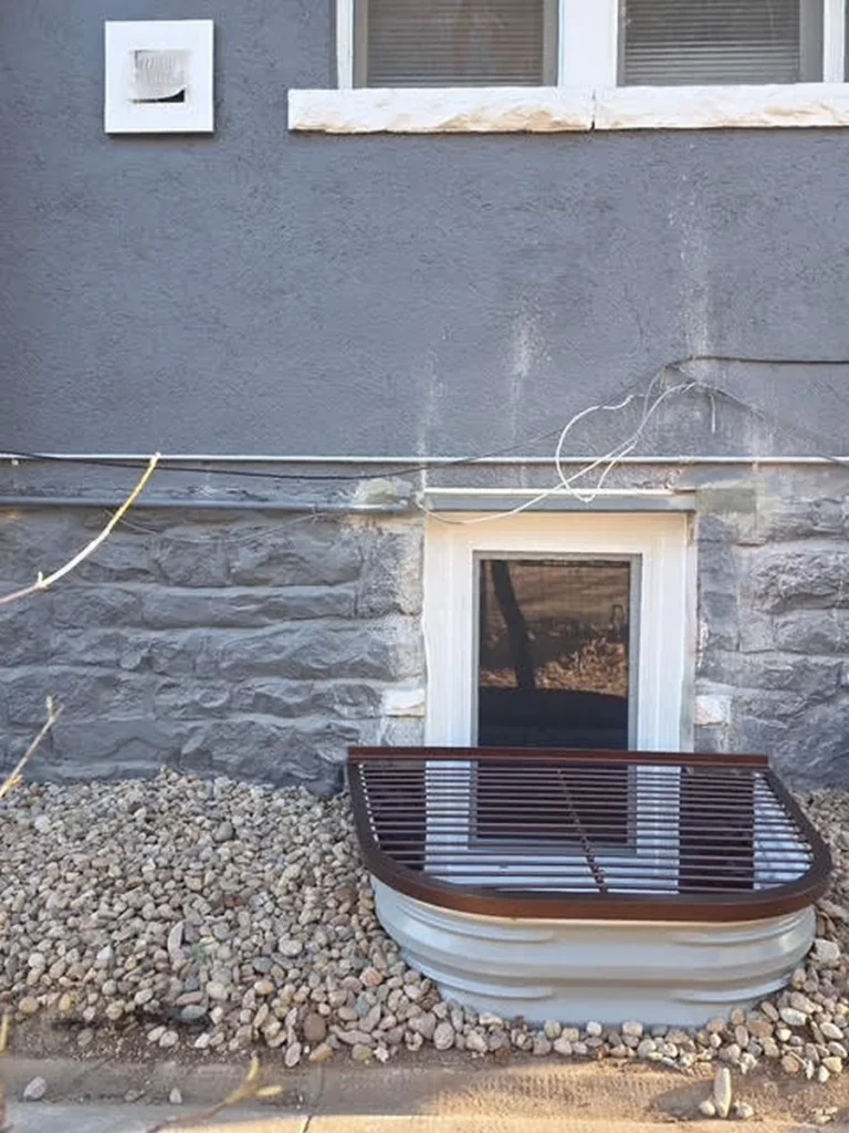 Stone foundation below a gray stucco wall beside a new egress window well and gravel